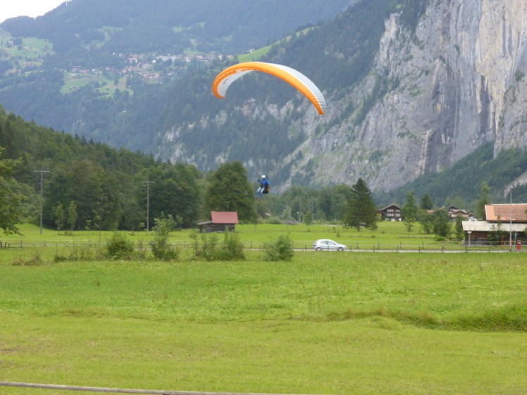 Watching a paraglider land as we wait for the rest of the group. It has an orange and white wing and is about to land in a field. In the background there are dark green pine trees and behind that, steep mountain cliffs. I think you can also see Murren up on its shelf on the left.
