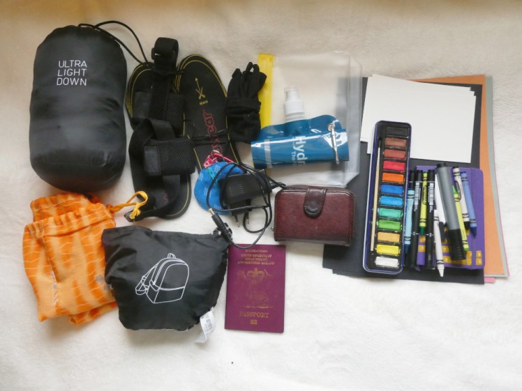 All my extras: a small bag containing a down jacket, a folded drawstring laundry bag, a pair of heated insoles with battery packs, a folding Ikea backpack, my passport, wallet, charger for the battery that keeps my coat warm, a blue round pouch for putting jewellery in, a folding flask and a silicon sandwich bag. To the right is a pile of sugar paper folded in half, some black paper and some A6 pieces of watercolour paper. On top is a child's tin of paints, a handful of pens, crayons and chalk and underneath, my notebook.