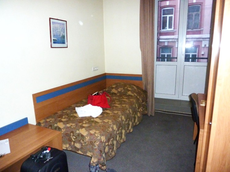My hotel room in Vilnius. It's dark - the carpet is a dark greyish blue, there's a kind of brown khaki-style cover on the bed, dark wood in the wall where the bed touches, a dark wardrobe right up by the camera and it's not all that light outside either.