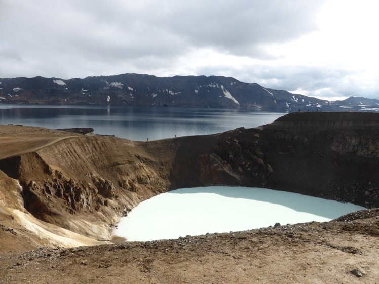 In the background, the deep blue Askja lake and the ring of mountains containing it. In the froreground is a smaller crater with muddy brown sides, flooded with opaque blue-white water that looks a bit like milk under the bright sunshine.