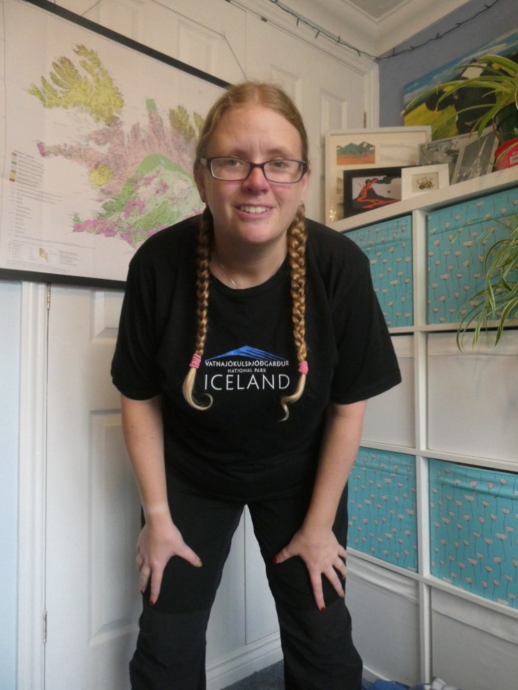 Leaning into the camera, wearing a black t-shirt with a national park logo on it. There's a stylised mountain in bright blue and underneath, the long name of the national park in small white letters. Underneath is ICELAND in bigger letters.