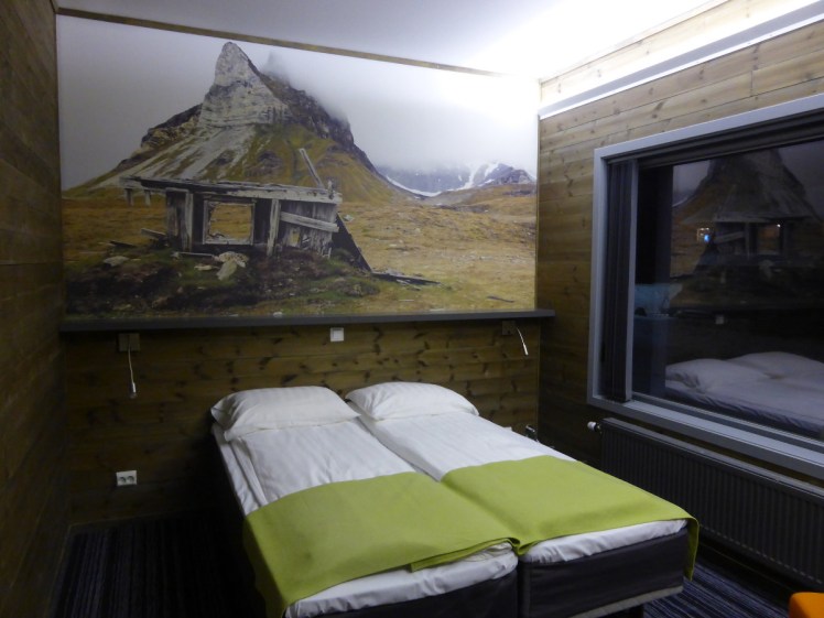 My hotel room in Svalbard. I have two single beds pushed together, there's a big picture of a mountain and some kind of tumbledown wooden structure dominating the wall above the bed and next to the bed is a huge window. It's so dark outside that all you can see is the reflection of the room in it.