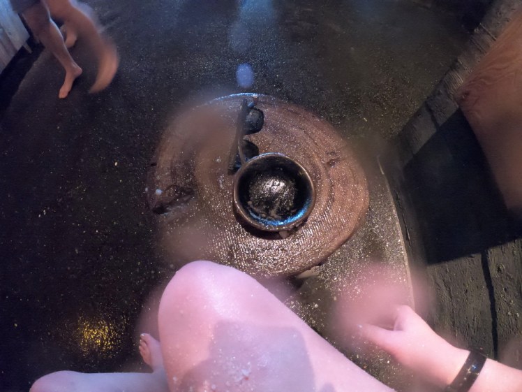 My leg and a little wooden bowl with the remains of a body scrub in it. I'm sitting in a darkened Ritual room and I'm using a log as a table for my bowl of scrub.