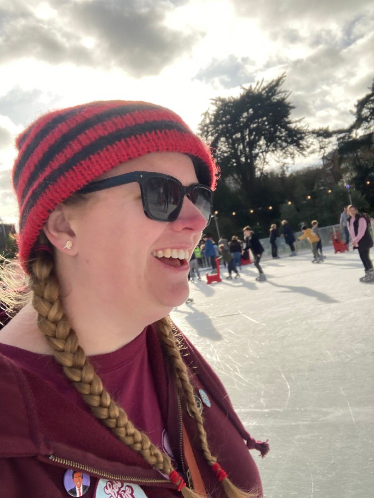 A selfie taken while ice skating. I'm wearing a red and black striped hat with a long tail and a burgundy t-shirt and hoodie.