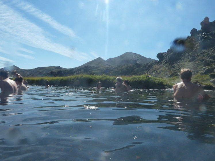 A few people sitting under a bright sun in the Landmannalaugar spring, which is a duckpond-sized shallow pool sparkling in the sun with a lava field rising up on the right and rhyolite mountains visible behind.