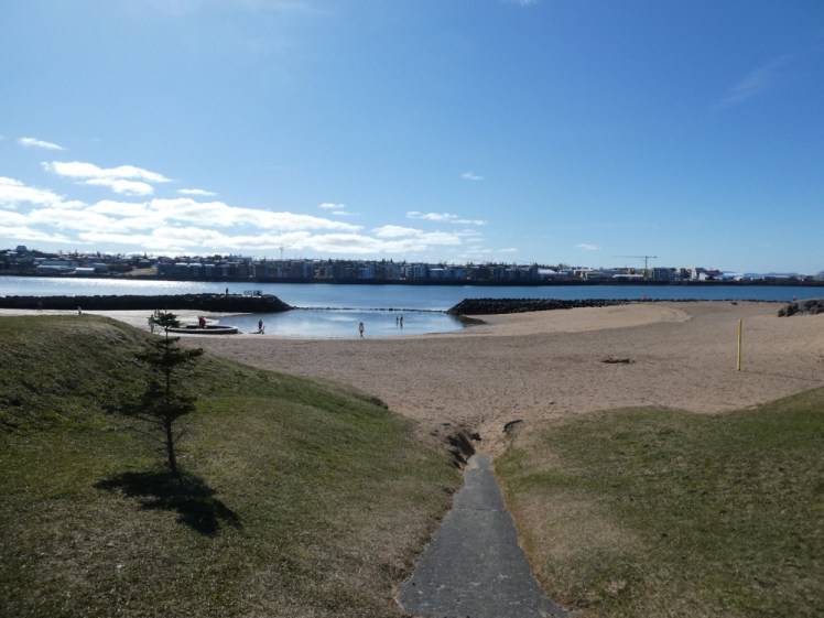 Nautholsvik as seen from the top of the path connecting it to the road, a dish of sand with a lot of blue water in the bottom. Separating beach from road is a bank of green stubbly grass.