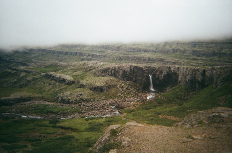 A waterfall in an ashy stratified cliffside, with grass clinging to it in layers. A heavy cloud blocks out the top of the mountain as if there's an error in the film (there isn't).