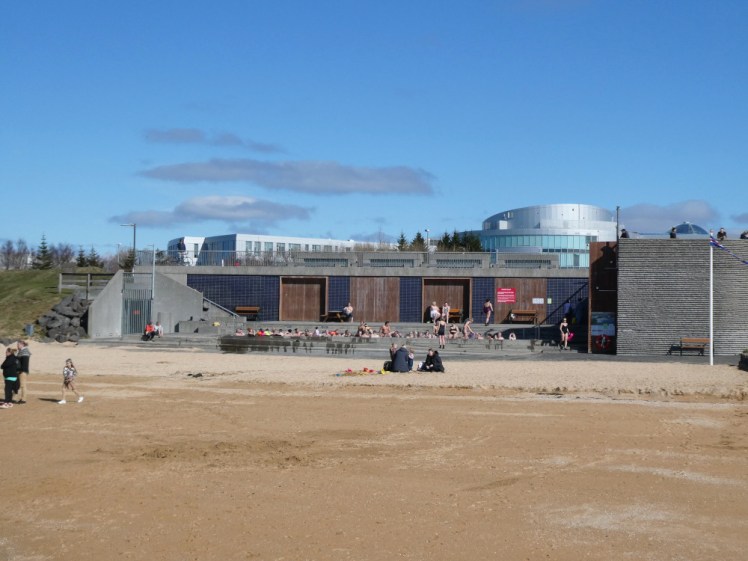 A concrete building set half into the grassy wall that forms the back wall of the beach. The front of the building is a series of long steps that has a long hotpot set into them.