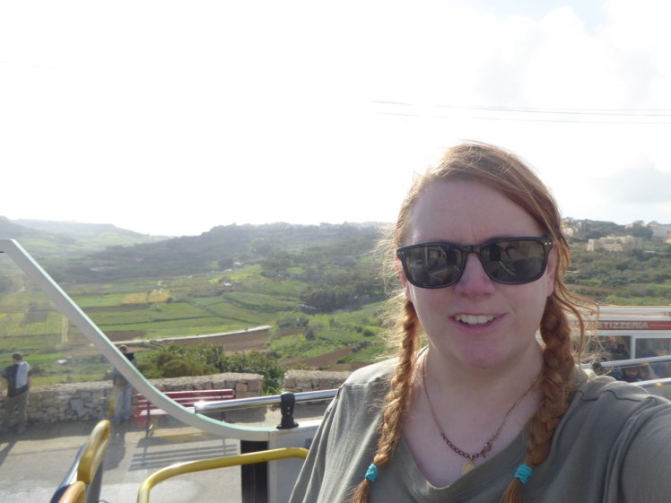 Me, with my hair in two plaits and looking very windswept, on the top deck of the tourist bus in Gozo. Behind me are green fields and hills. It's very hot and it's only January.