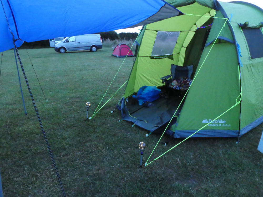 A large green tent with an open front. Attached to it is a blue tarp. It's just a little dark although what little you can see of the sky is still white.