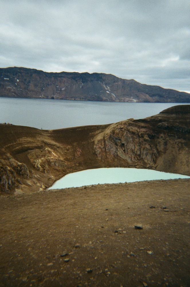 A pool of milky blue water in a muddy crater. Behind it is a larger lake with bluer water and a ridge of mountains on its opposite shore.