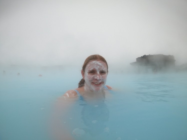 A selfie in the Blue Lagoon. It's very misty behind me, with no clear division between sky and water. I'm in the middle, arm extended towards the camera with a silica mud mask on my face.