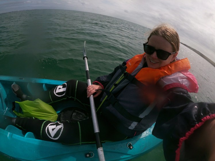 Me in a blue sit-on-top kayak out at sea, wearing a wetsuit that you can hardly see under my pink and black jacket and my orange buoyancy aid. 