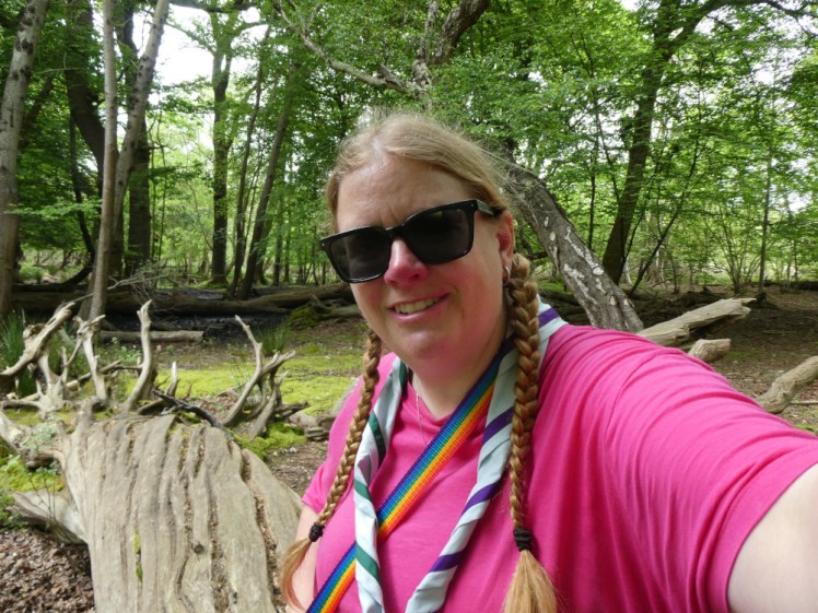 Me, in a bright pink t-shirt and wearing the Brownsea neckerchief, which is pale blue with dark green and purple borders on either side, sitting on a fallen tree in woodland.