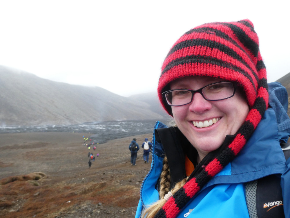 Me, wearing a red and black striped hat with a long tail, grinning very happily at the camera with a black lava flow behind me. The lava is steaming and the cloud is low.