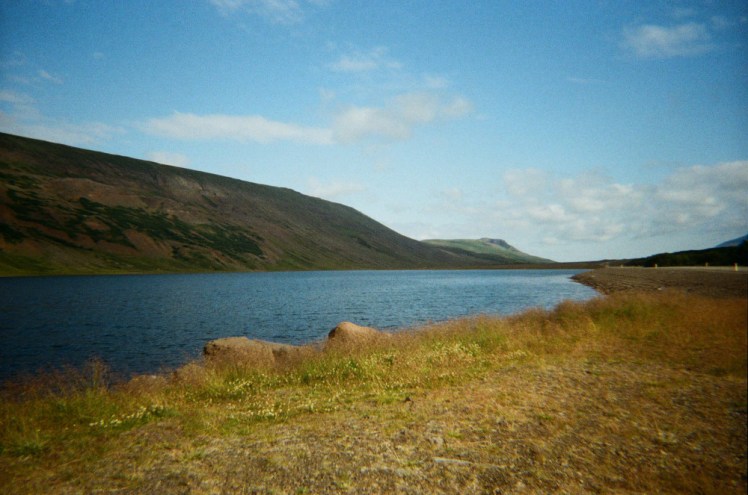 A very blue lake between a long low mountain and a scrubby grassy shore. The sky above is blue.