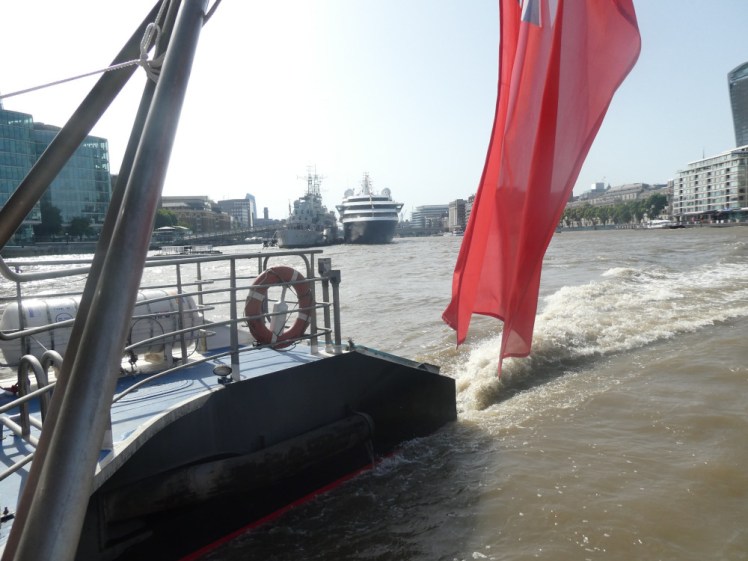 The view from the back of the Clipper. The Thames is brown and well-churned by our engines and behind us is a cruise ship moored so that it obscures HMS Belfast from the point of view of a boat whooshing past them.