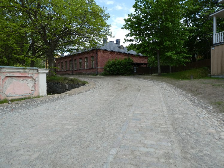 A large but single-storey red brick building at the bend in a cobbled road. There are bright green trees almost touching on each side of the road.