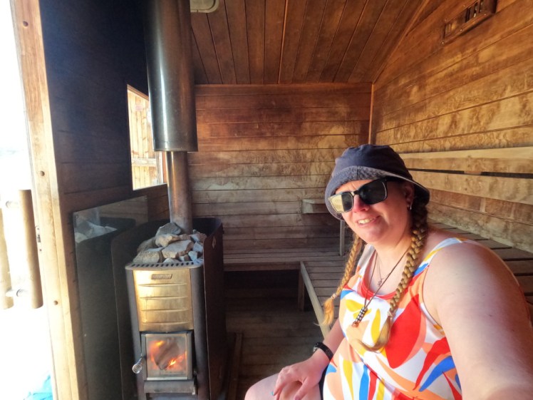 Me in a bright-coloured swimsuit sitting inside the sauna hut, which looks like a wooden shed with a wood burner in it.