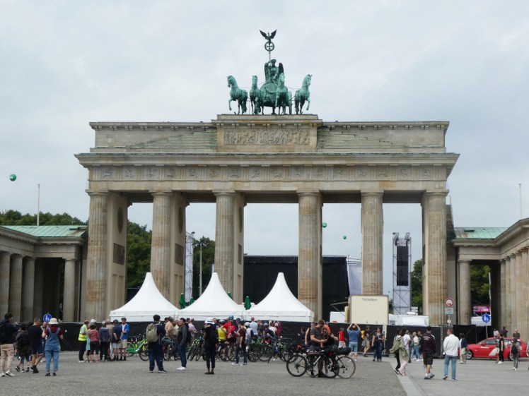 Brandenburg Gate, a large stone classical-style gateway topped with a green statue of four horses pulling a carriage. Below it are large gazebos, a toilet van, blacked-out fences and lots of people trying to take pictures over the top. Behind it you can see the back of a stage and festival lighting.