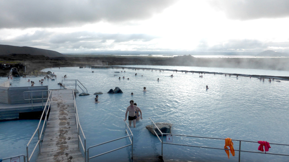 Myvatn Nature Baths. A vast blue lagoon with underwater steps stretches out. Beyond, you can see a lake shining on the horizon and mountains beyond even that.