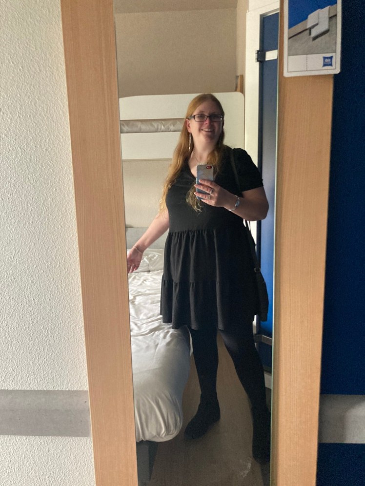 A really bad mirror selfie in a pretty bad hotel. I'm wearing a short-sleeved black dress with a fairly short skirt that flounces out a little, black tights and boots and for once, I have my hair down.