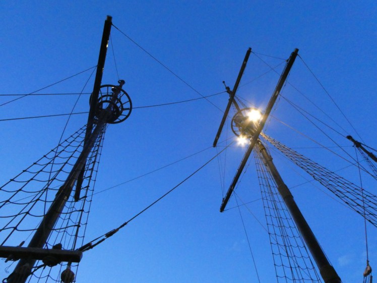 Masts and their rigging seen from below, silhouetted against the blue evening sky.