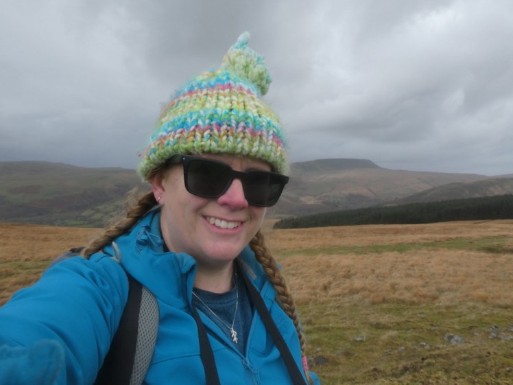 A selfie, wearing a blue softshell and a bright stripy bobble hat. I'm on the moors in Wales, with Fan Breichiniog behind me, on a wet and wild-looking day with swirling dark grey clouds.