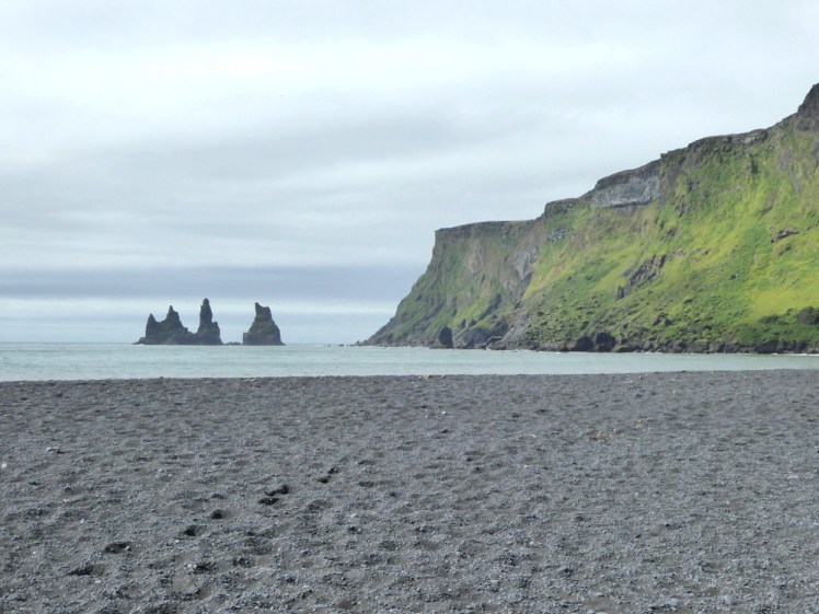The black sand beach at Vik. The sand is actually a dark grey, as are several jagged pinnacles sticking 70 metres out of the sea. To the right is a rocky headland, its steep slopes covered in luminous green grass and moss.