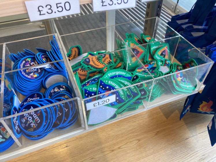 Boxes of badges at the Guide shop. To the left are round navy badges with bright blue edges, for Marshmallow Toaster and Teamwork. To the right are large green-edged badges in interesting shapes for various outdoorsy things like Stargazer and Trailblazer.