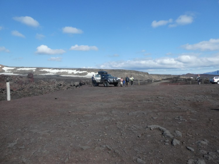 The Askja car park, a patch of slightly reddish gravel, surrounded by very rough dark grey lava. Behind it, a mountain ridge speckled with snow hems in the lava.