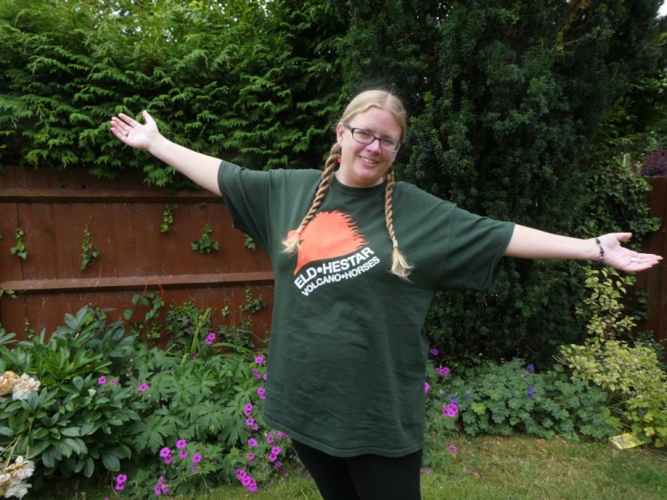 Me, in the garden with my hair in plaits and arms outspread, wearing an oversized dark green t-shirt. It has an orange horse's head on it and underneath says Eld.Hestar, volcano.horse.