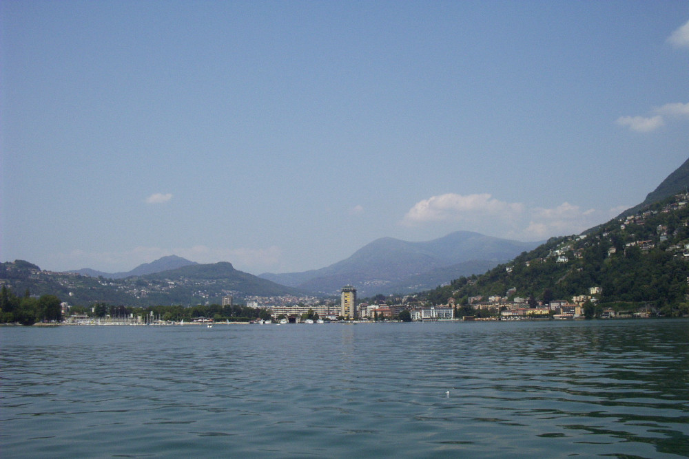 On the far side of a lake is a town. To the right of the town, a mountain rises up steeply and there are more mountains in the hazy distance. All the mountains are pretty green and dotted with buildings.