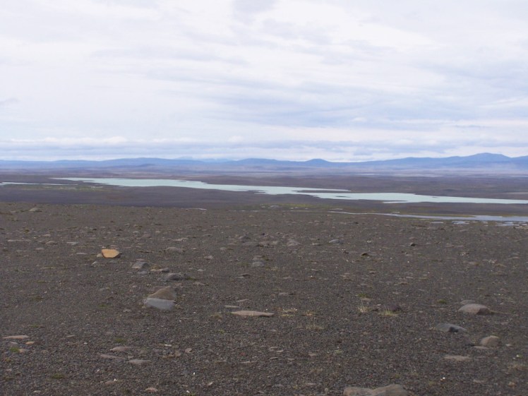 A view of the Icelandic Highlands, a huge expanse of brown gravel with no visible roads or tracks, a wide river meandering around the bottom and hazy blue snow-tipped mountains in the distance.