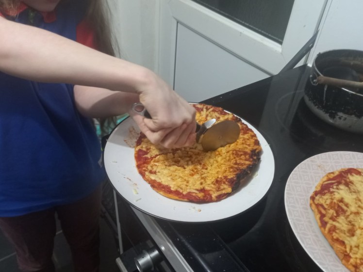 Overcooked pizza burned around the edges, being cut up by an unseen Guide. You can just see a glimpse of blue and red uniform.