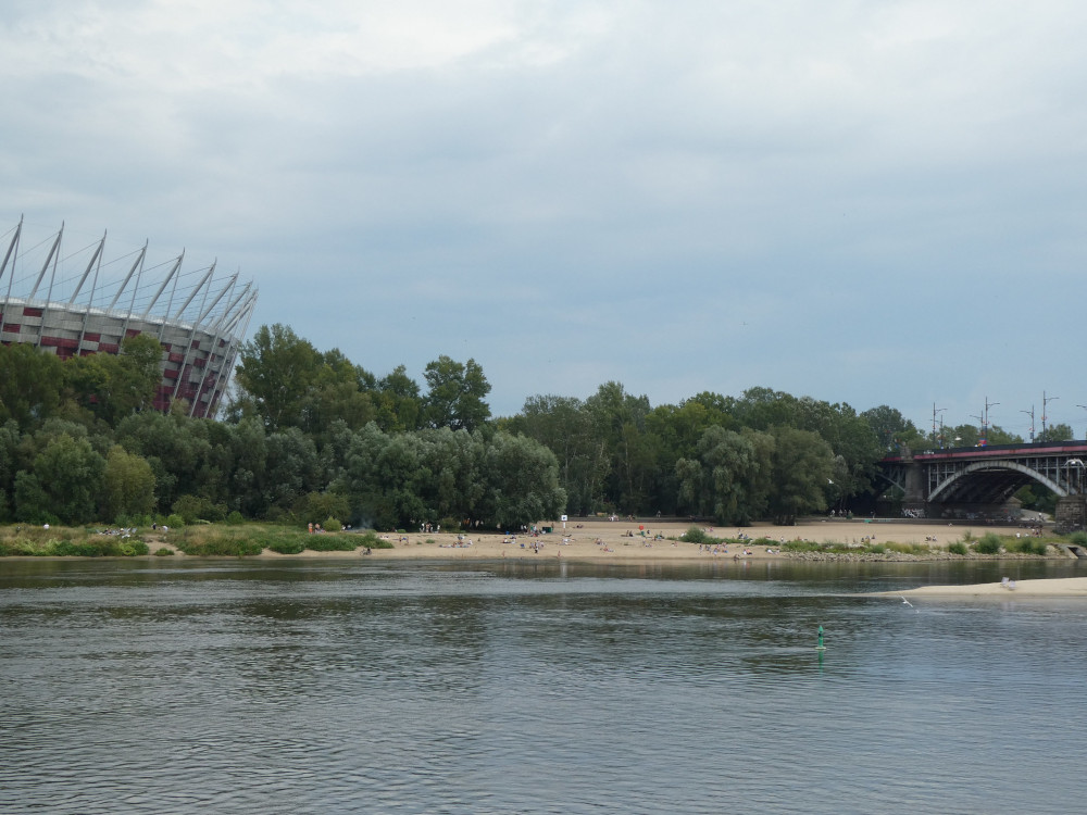 Walking along the river. The other bank is all trees with the national stadium rising out of it on the left and an area of sandy beach towards the right.