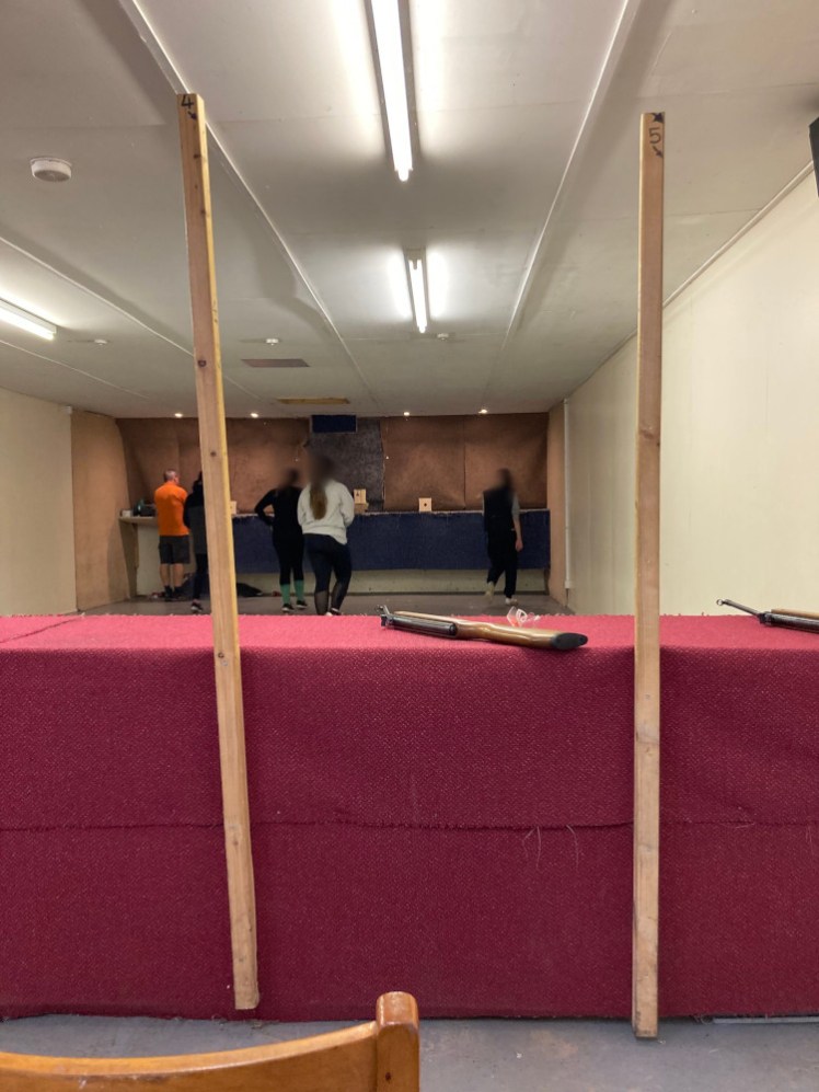 The rifle range, a long room viewed from behind a table separated by upright wooden batons. My Rangers - blurred but with their backs to the camera anyway - are collecting their targets.