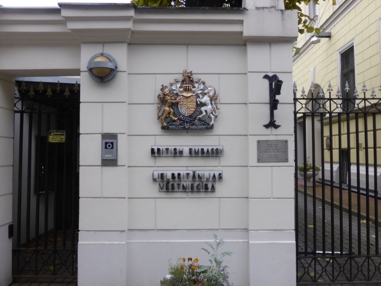 The British Embassy in Latvia. A white stone wall with the British coat of arms. To the left is a dark passageway with a gate across it. To the right, a path between it and the next building is also closed by a gate.