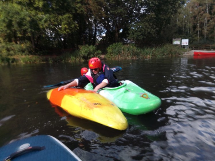 Me in a tiny green kayak leaning over to try to right un upturned yellow kayak in a river.