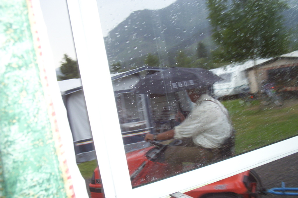 A blurry photo of Wolfgang, the campsite owner, driving his little train pulled by a ride-on lawnmower. It's raining and he's holding an umbrella in one hand as he drives.