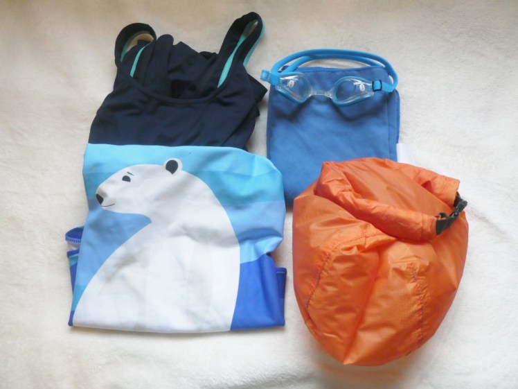 A navy swimsuit with teal edging and a blue swimsuit with a polar bear on it, folded up next to a blue travel towel and a bright orange drybag. On top of the towel is a pair of blue goggles that are so badly misted and frosted that it's time I got new ones. They're prescription and you can see the number on a sticker on each side.