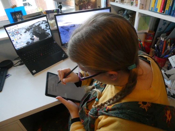 The camera is positioned above my head looking down at me sitting at my desk. I'm holding my tablet in my hands and writing on its screen with the S-pen, which looks like a slim black pen. I'm wearing a yellow jumper under the green dungarees you'll have seen several times lately and my hair is plaited.