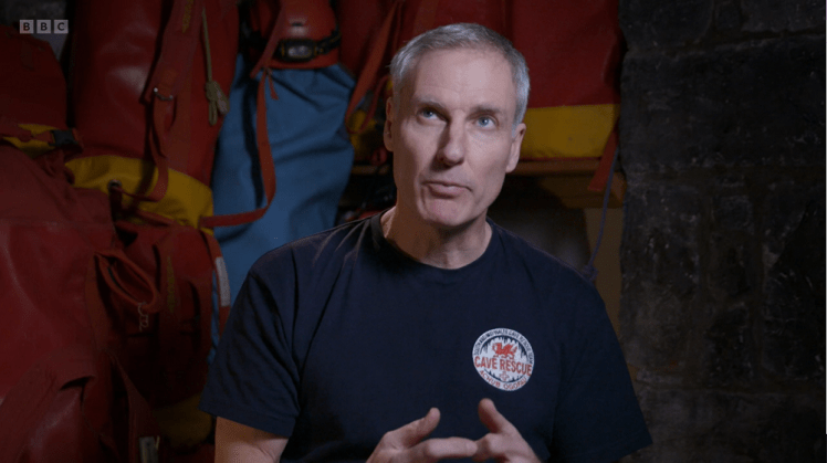 Gary Evans from cave rescue. He's a grey-haired man with a vaguely military bearing, wearing a navy t-shirt with the cave rescue logo on it. He's in the same store as George earlier, surrounded by red bags and blue and red caving suits.