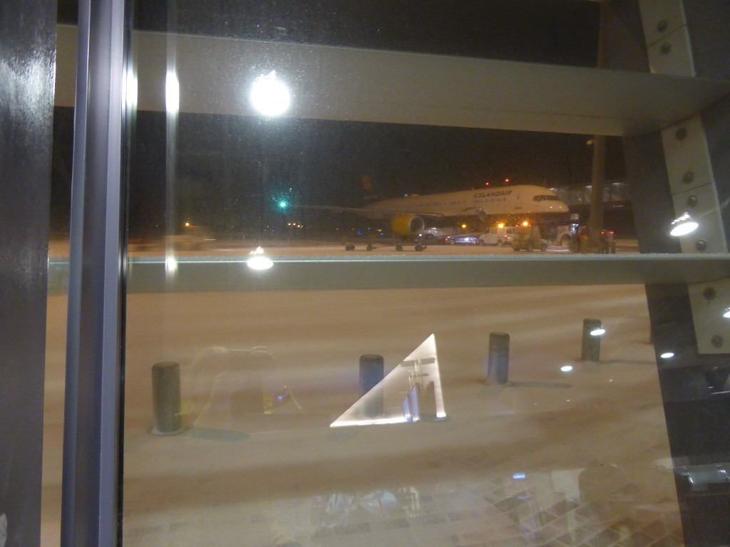 I'm sitting at a table in Keflavik Airport charging my phone. It's dark and snowy outside, with reflections on the window and a plane just visible.
