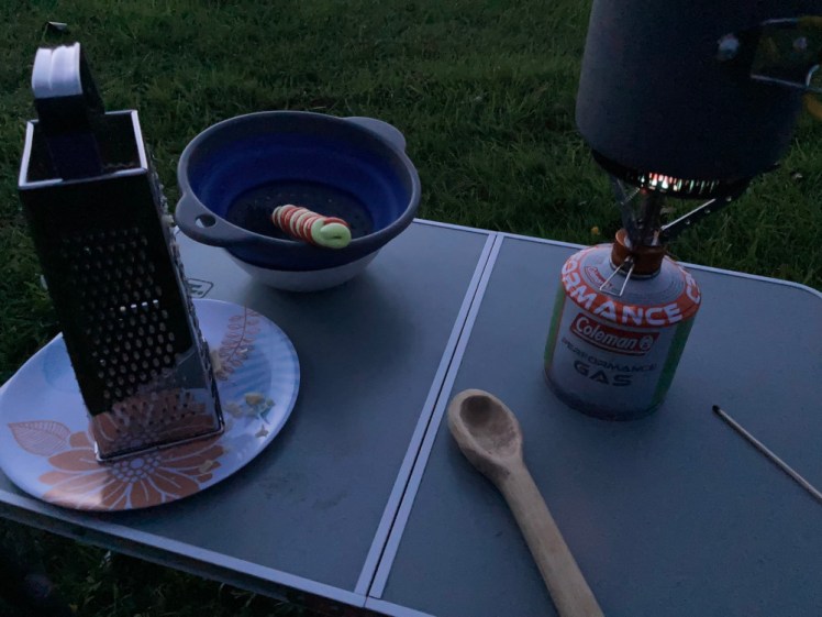 My tiny camp table outside in the dim light of evening. On the table is a cheese grater on a plate, a collapsible colander containing a camp spoon wrapped in orange and glow-in-the-dark Fimo, glowing slightly, a hand-whittled wooden spoon and a pot balanced on a gas stove.