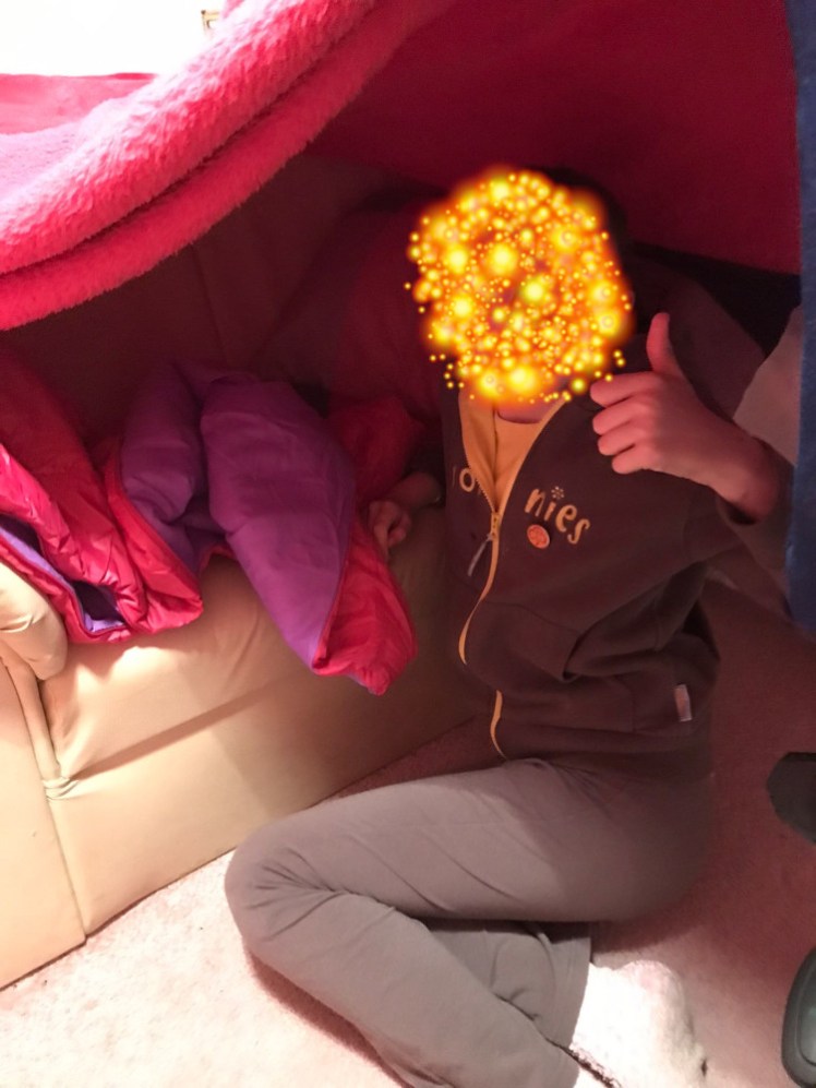 A Brownie in her den, with a huge supernova covering her face for anonymity. The den is a pink blanket roof over lots of pink blankets and a girl in Brownie uniform holding her thumb up to the camera.