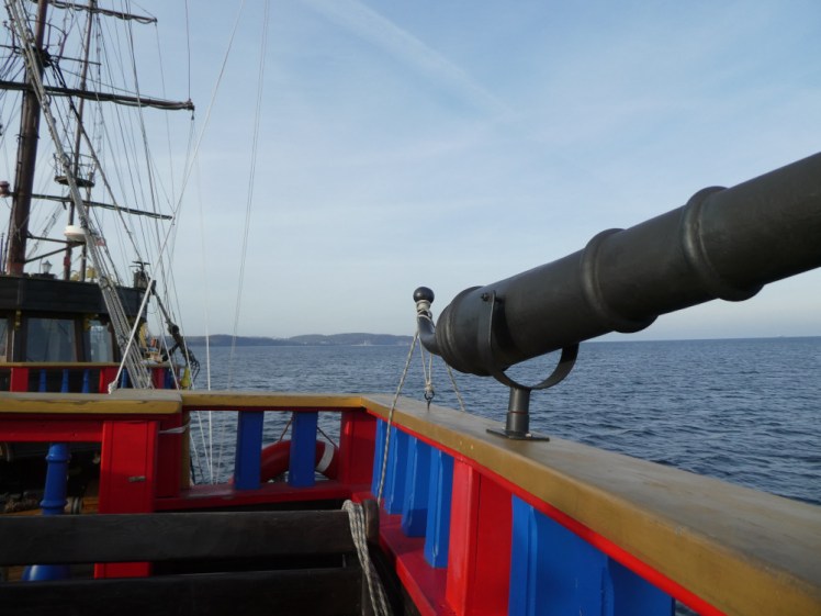 Looking forward from a rear deck of the pirate ship with a good amount of sea on the right. The railings are bright red and blue and the rail around the top is gold. There's a toy cannon or maybe telescope bolted to the rail which doesn't add to the realism of it all.