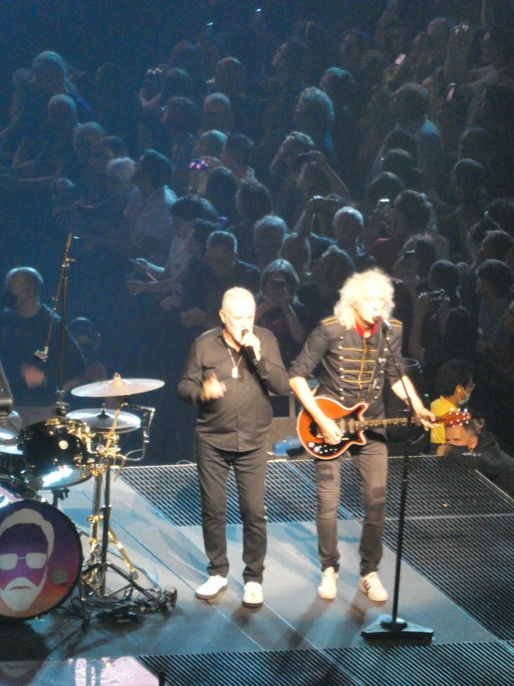 Roger Taylor and Brian May performing together. Roger Taylor is wearing a black tracksuit and Brian May is still in his black jeans and a military jacket with a red collar. Taylor is singing and May is playing an electric guitar.