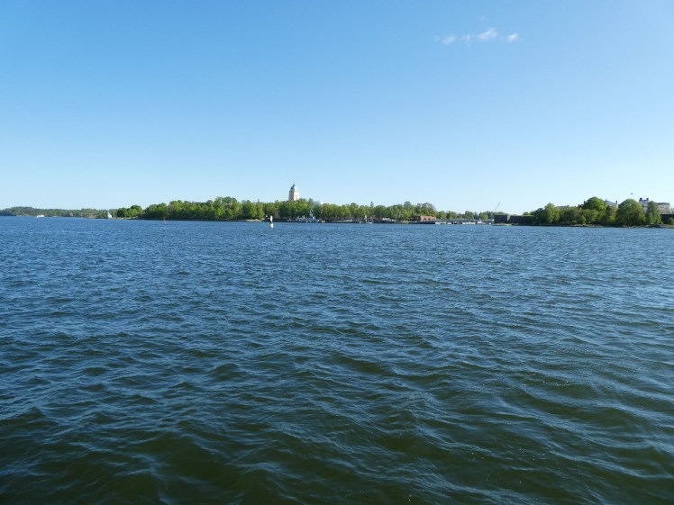 An island of trees with a square church spire sticking out of it. The water is green-blue and the sky is perfect and blue.
