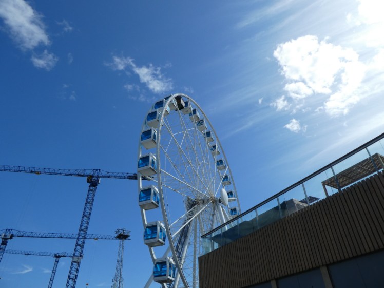 A large ferris wheel with blue-windowed cabin seen from below. Behind it are cranes and in front is the corner of Allas Sea Pool's raised wooden gallery/bar.
